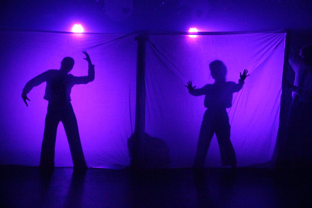 Bag murene: to dansere bag stofvæg, hvor de laver skyggedans i lilla lys.